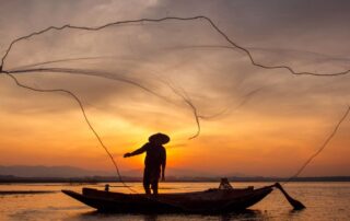 A stunning silhouette of a Vietnamese fisherman casting a traditional net on the river at sunset, representing the authentic culture of the Hoi An and Da Nang region.